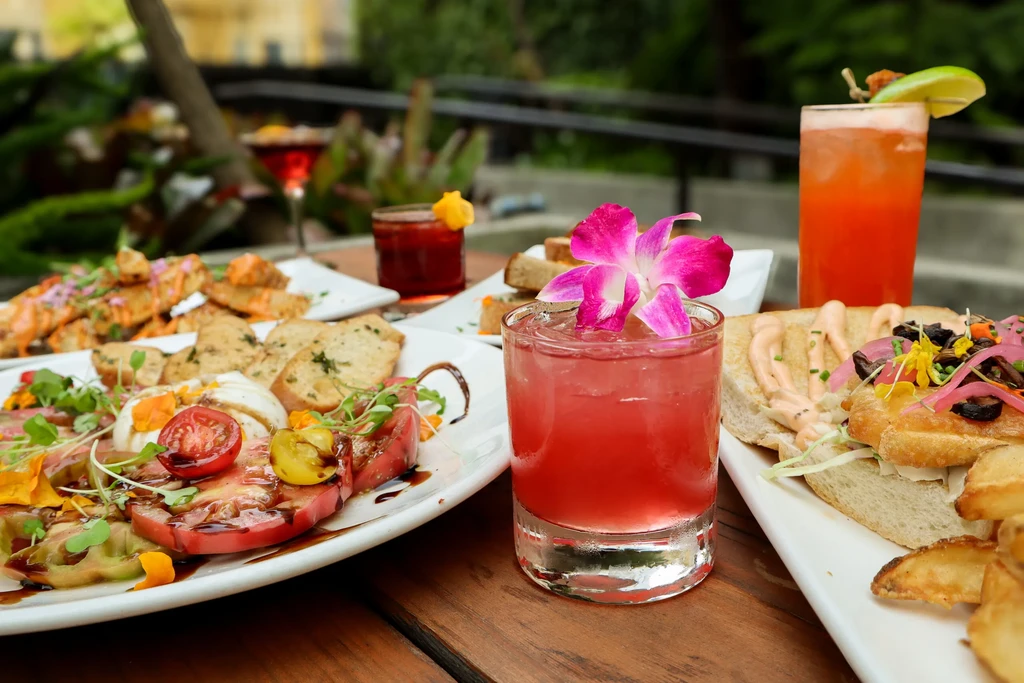 Several colorful cocktails and plates of food on a wooden table.