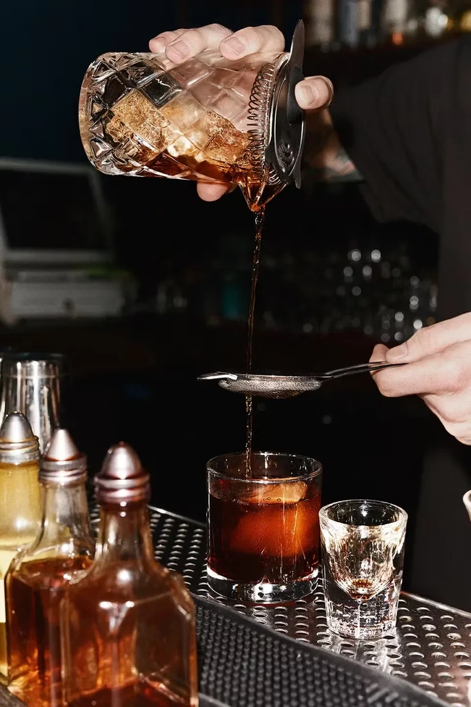 A bartender strains an amber cocktail into a glass with ice.
