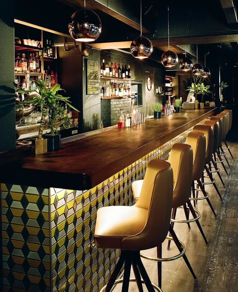 A dark, modern bar with a long wooden counter and yellow stools.