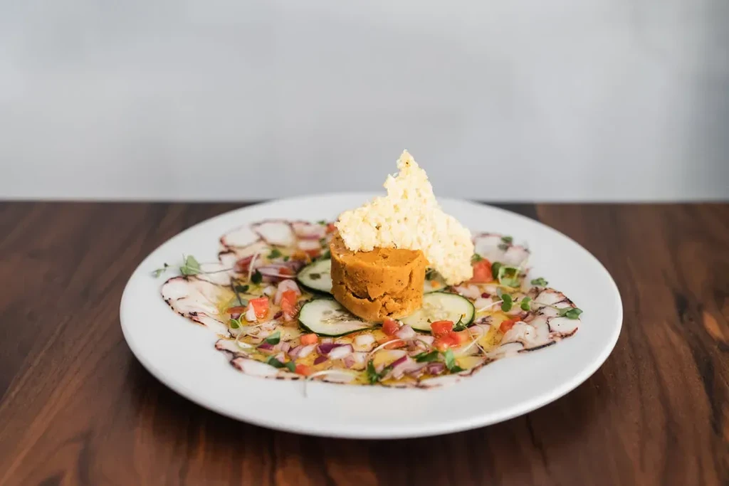 Plate of octopus carpaccio with vegetables, a pâté-like tower, and cheese crisps.