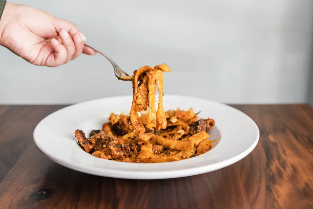 A fork lifts pasta from a white plate on a wooden table.
