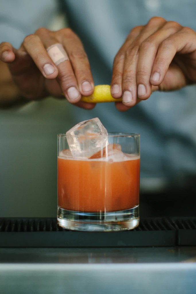Hands squeezing a lemon peel over a glass with an orange cocktail.