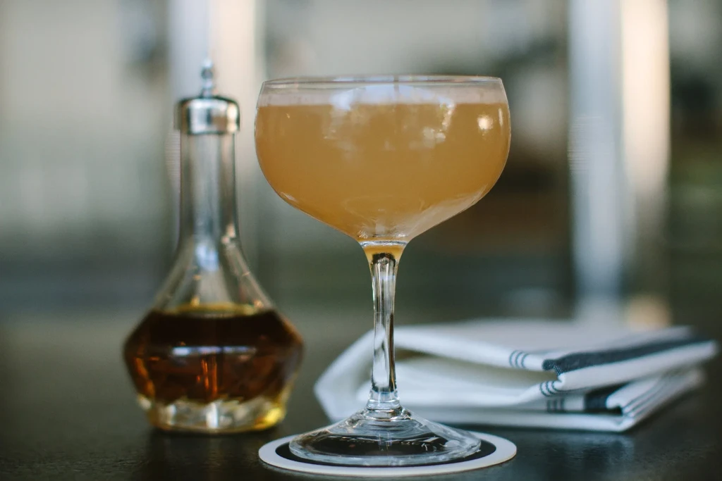 A coupe glass with a light brown cocktail next to a small bottle.
