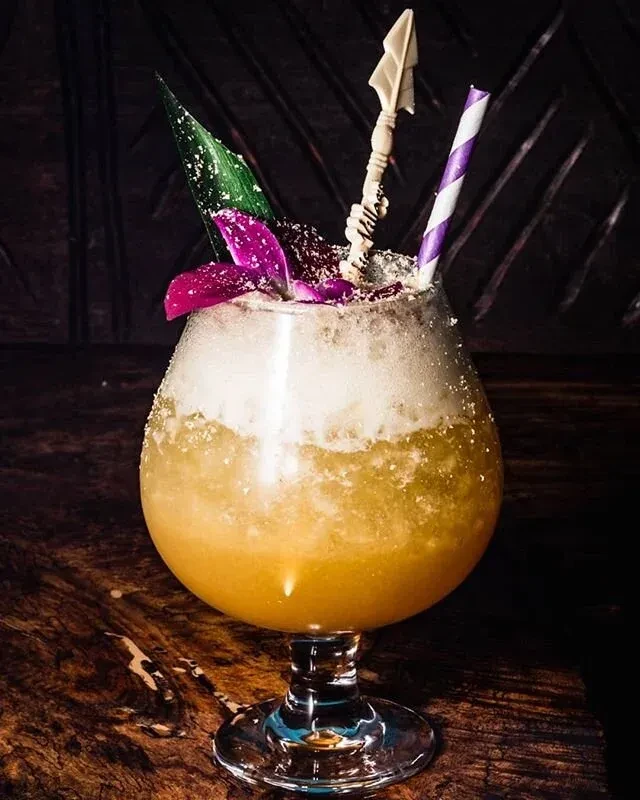 A colorful cocktail with a tropical flower and arrow stirrer on a wooden surface.