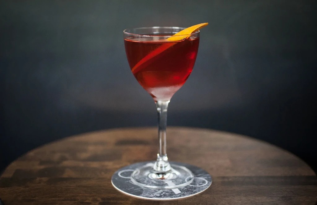 A red cocktail in a stemmed glass with an orange peel garnish.