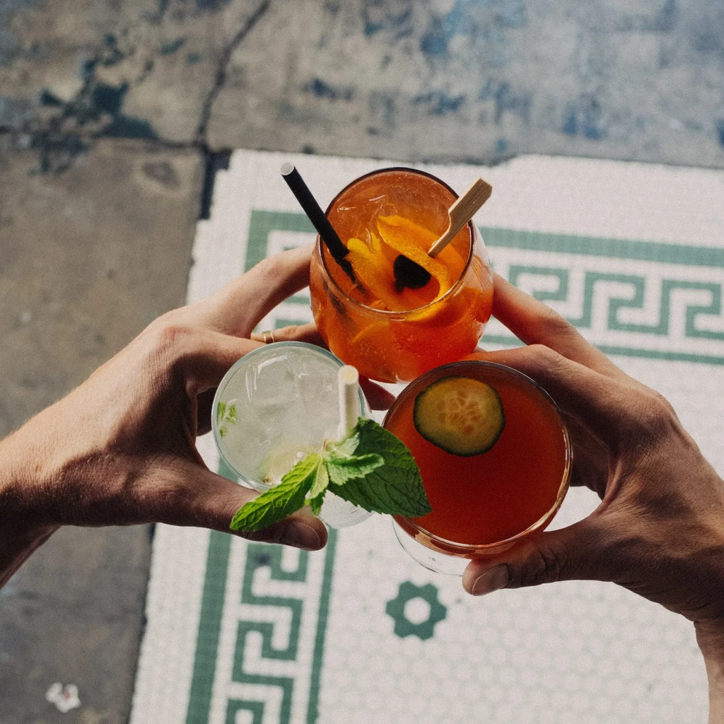 Three hands hold up three cocktails with different garnishes over a tiled floor.