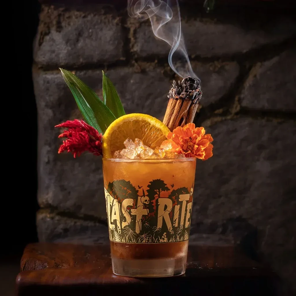 A cocktail in a "LAST RITE" glass with garnish and smoking cinnamon.