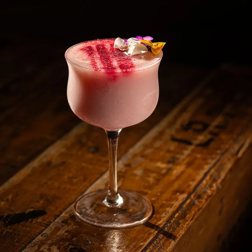 A pink cocktail in a stemmed glass with floral garnish on a wooden surface.