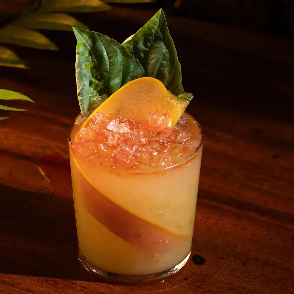 A cocktail with crushed ice, orange slice, and green leaves on a wooden table.