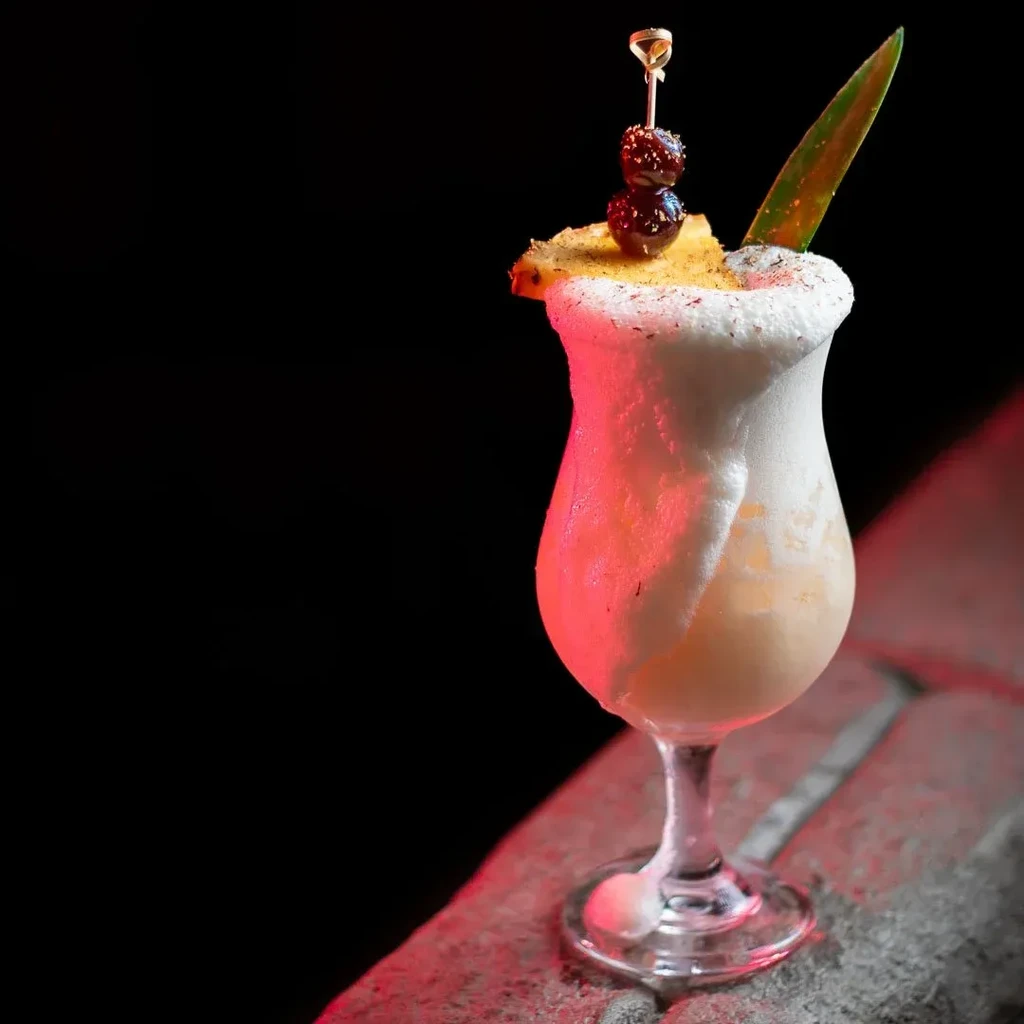 A tall cocktail with frothy white foam, garnished with pineapple and cherries.