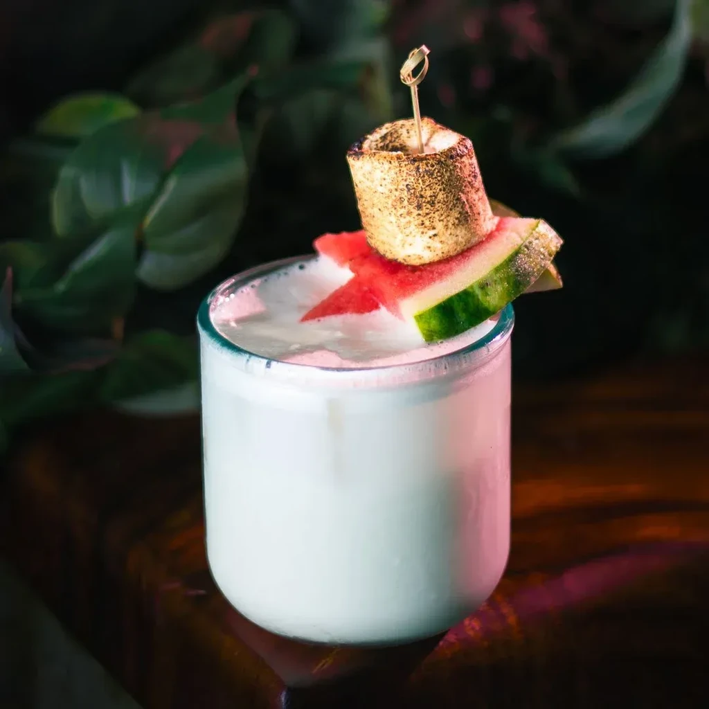 A white cocktail in a glass, garnished with watermelon and a toasted marshmallow.
