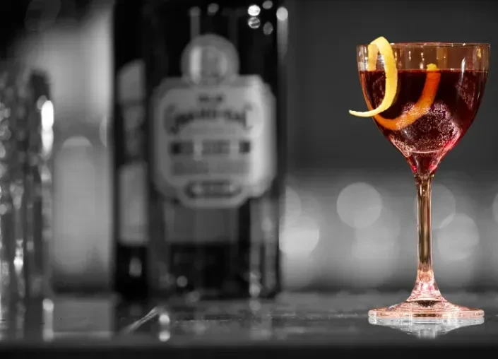 Red cocktail with lemon peel garnish, bottle in background.