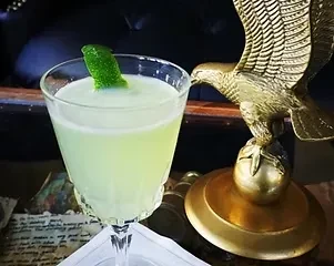 A pale green cocktail with a lime wedge, next to a golden eagle statue.