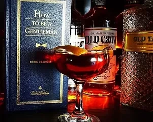 A dark red cocktail next to a book titled "How to be a Gentleman."