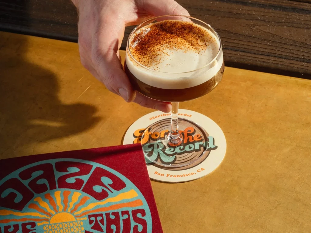 Hand holding cocktail with foam and spice, on "For the Record" coaster.