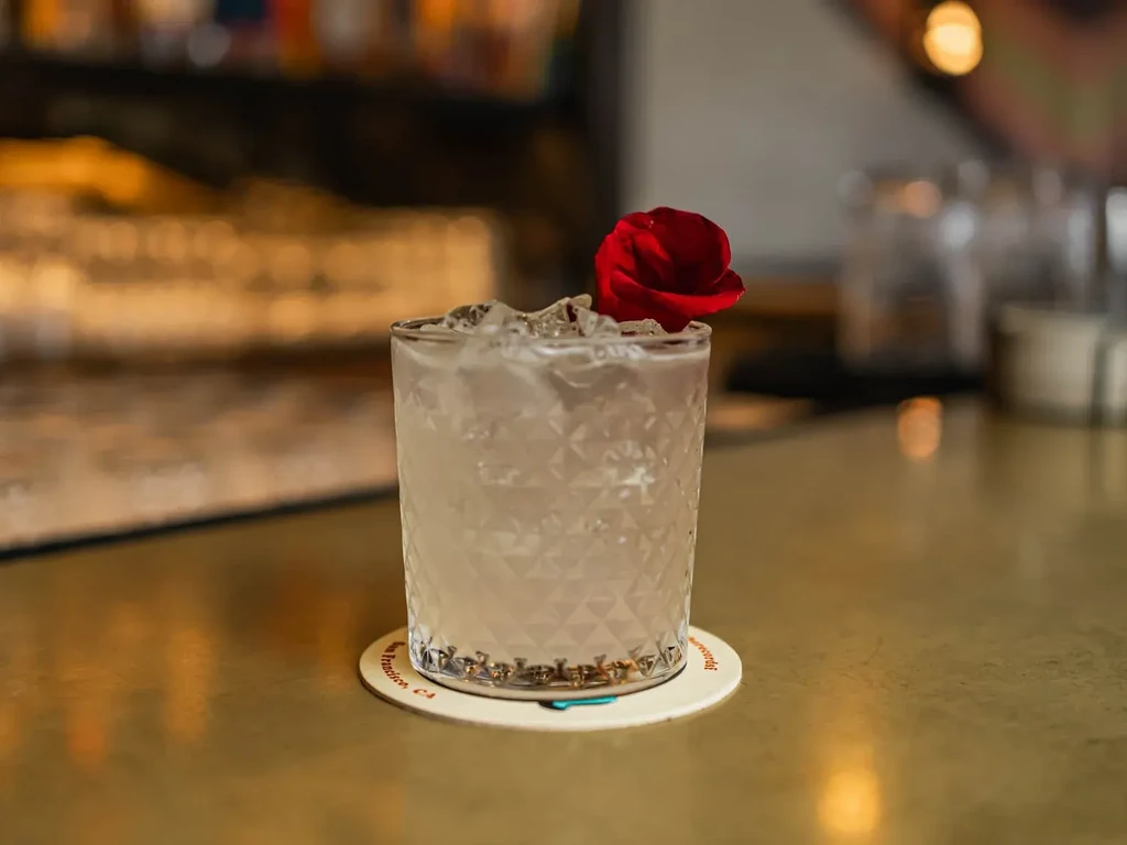 A clear cocktail with ice and a red rose petal on a coaster.