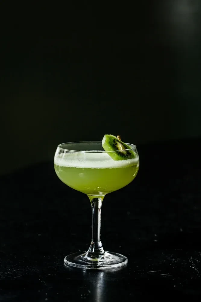 A green cocktail in a coupe glass with a kiwi garnish.