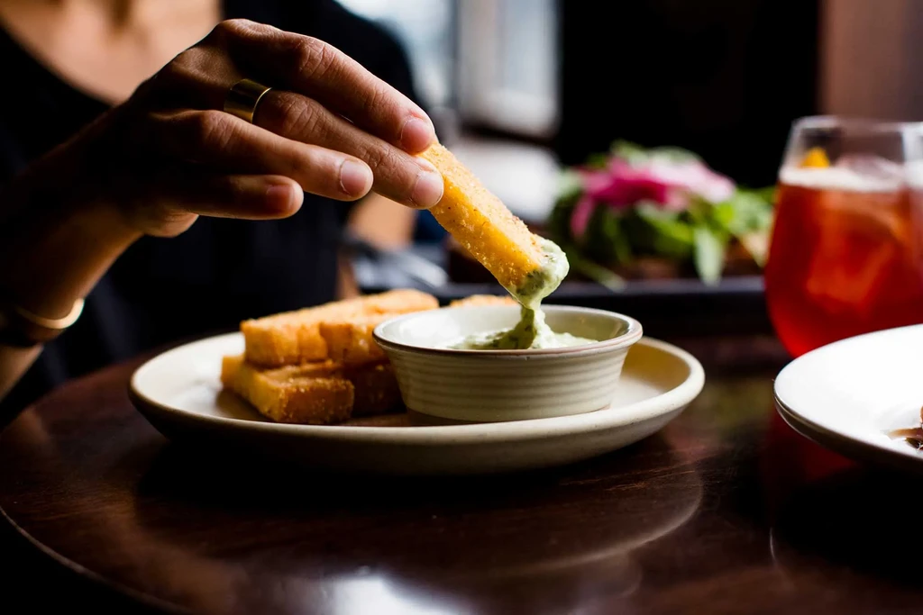 Hand dipping a golden fried polenta stick into green sauce.