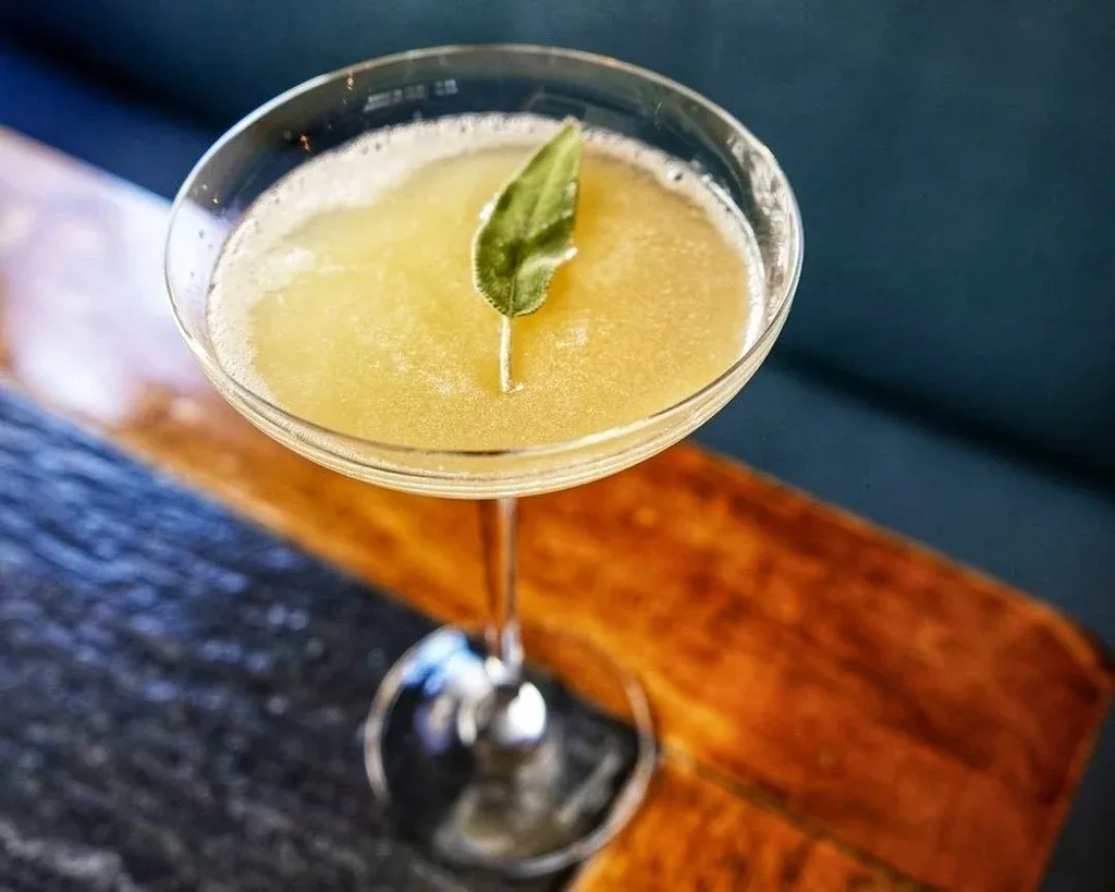 A pale yellow cocktail in a martini glass garnished with a leaf.