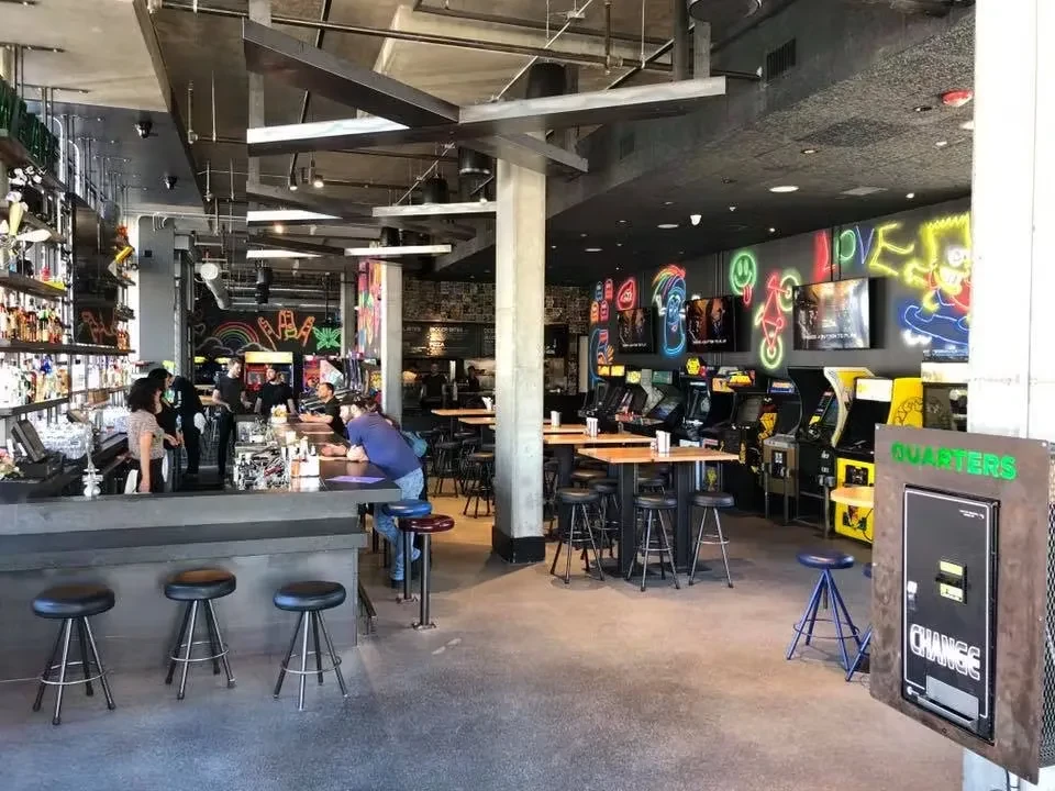A bar with arcade games, neon signs, and people socializing.