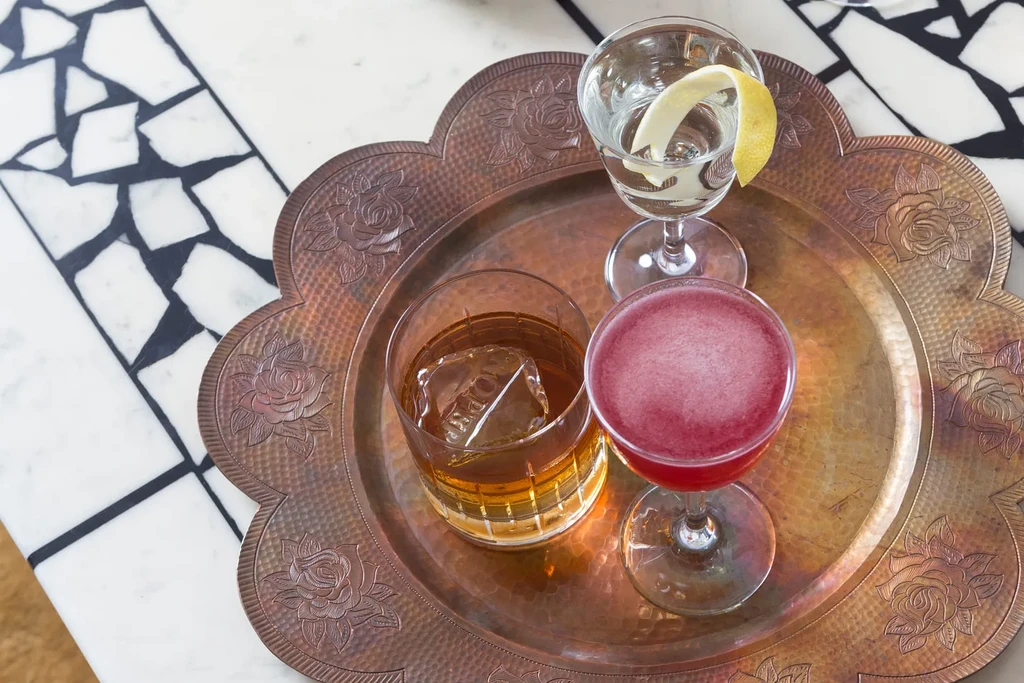 Three cocktails on a rose-patterned copper tray, on a white mosaic table.