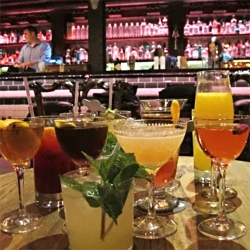 Various cocktails sit on a table in front of a busy bar.