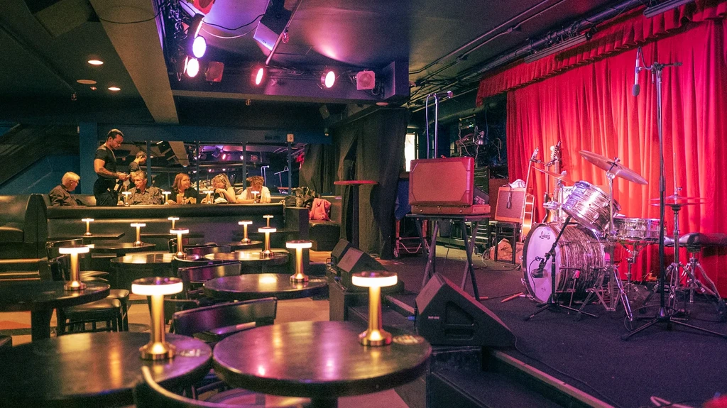 A bar with tables, patrons, and a stage with a drum set.