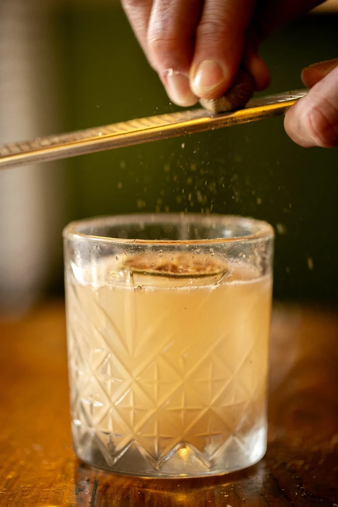 Hands grate nutmeg over a light-colored cocktail in a crystal glass.