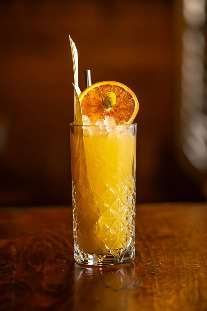 A tall yellow cocktail with ice, dried orange, and a leaf garnish.