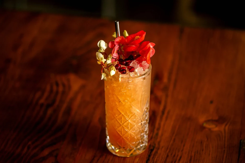 A tall cocktail with crushed ice, fruit, and red flowers on a wooden table.
