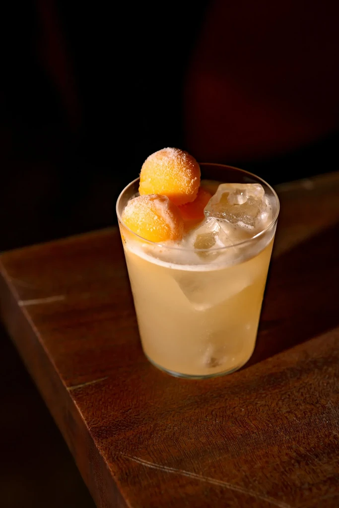 A clear glass with a light yellow drink, ice, and three frozen melon balls.
