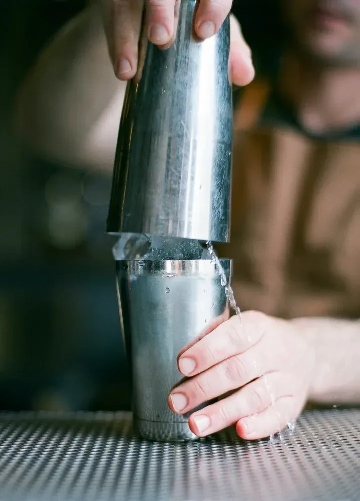 A bartender holding a cocktail shaker, water spilling onto a metal surface.