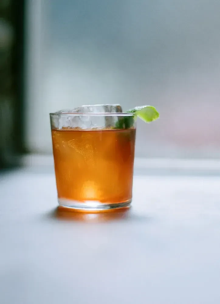 A glass of amber cocktail with ice and a lime peel garnish.