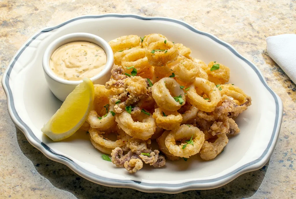 Plate of golden-brown fried calamari with a lemon wedge and dipping sauce.