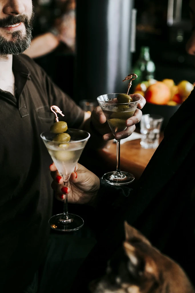 Two people hold martini glasses garnished with olives at a bar.