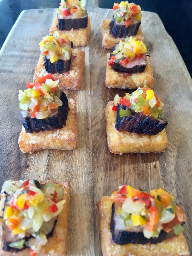 Seven appetizers with meat and salsa on potato squares.