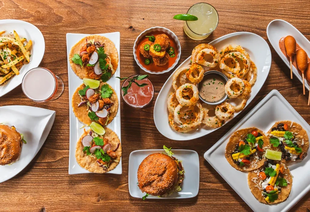 A table filled with various dishes: tacos, burgers, onion rings, and cocktails.