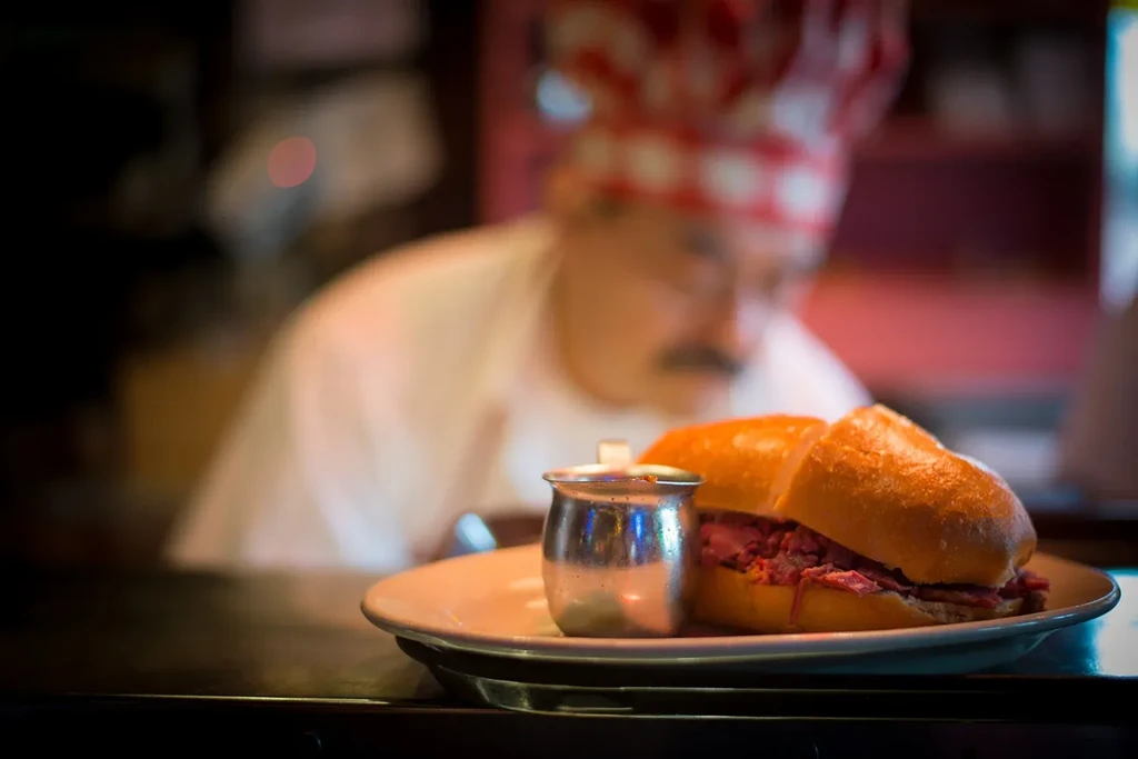 Chef in white uniform behind a plate with a sandwich and sauce.