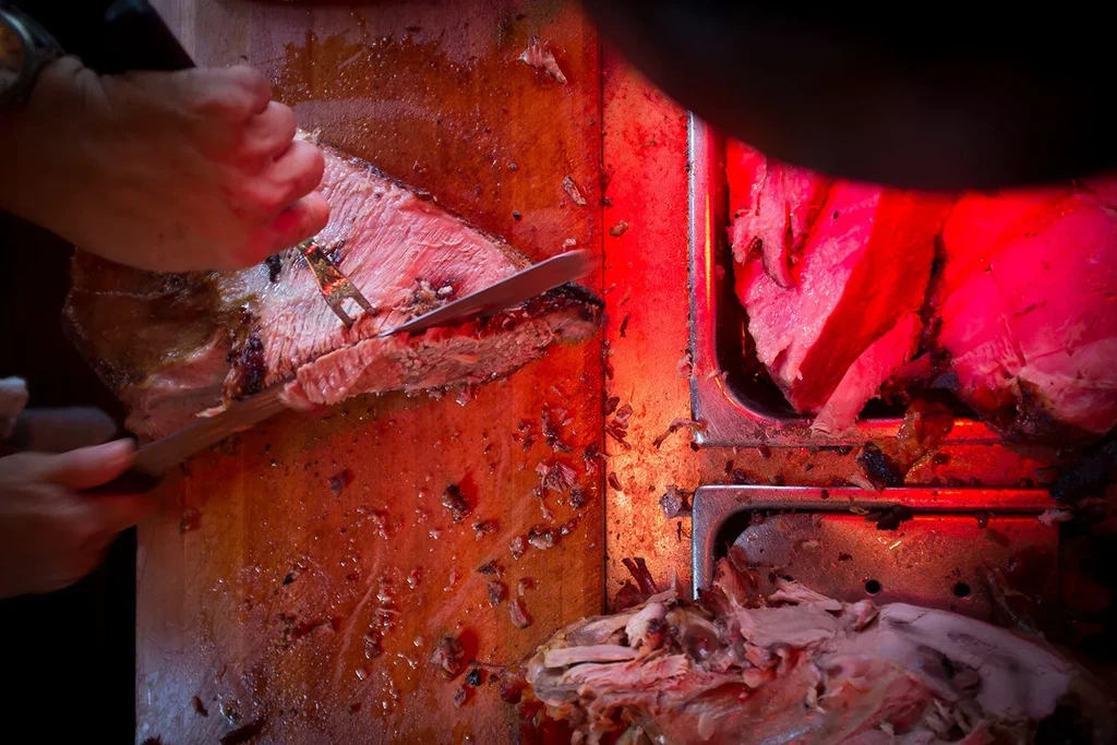 A person carving a large piece of meat under a red light.