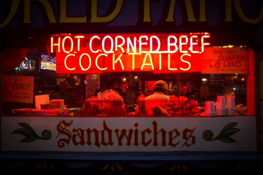 Neon sign for "Hot Corned Beef Sandwiches" and "Cocktails" at night.