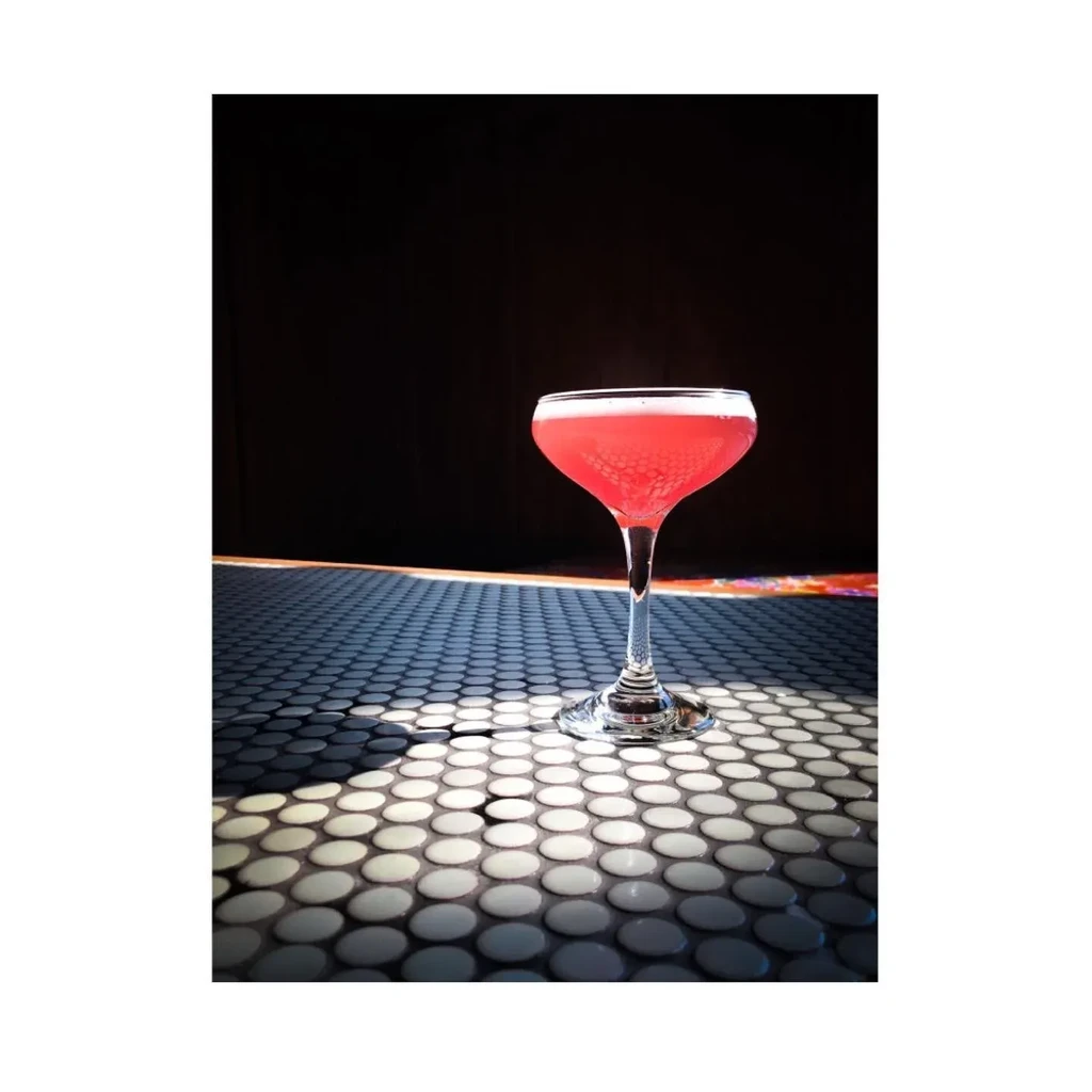 Pink cocktail in a coupe glass on a white tiled surface.