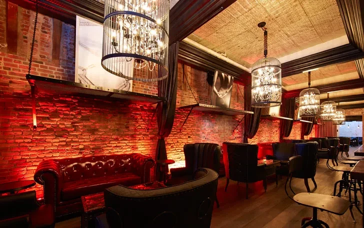 Interior of a dimly lit bar with red lighting and caged chandeliers.