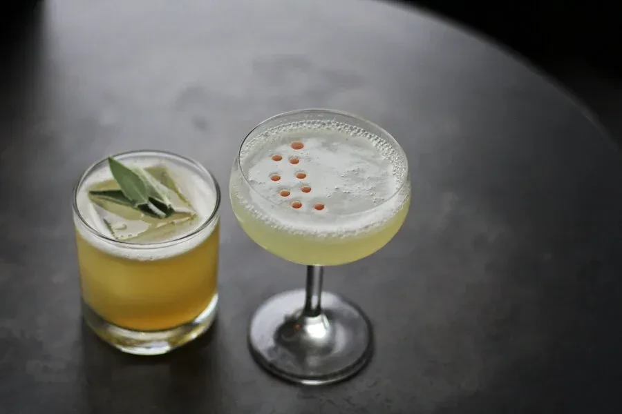 Two yellow cocktails, one garnished with sage, on a dark table.