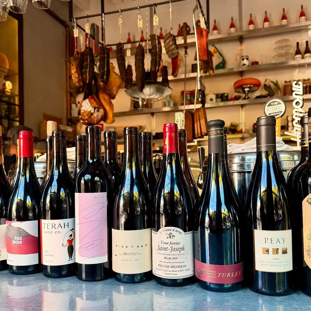 Wine bottles on a counter in front of hanging cured meats.