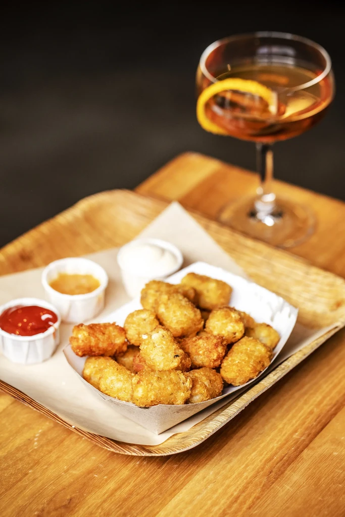 Tater tots with dipping sauces and a cocktail on a wooden tray.