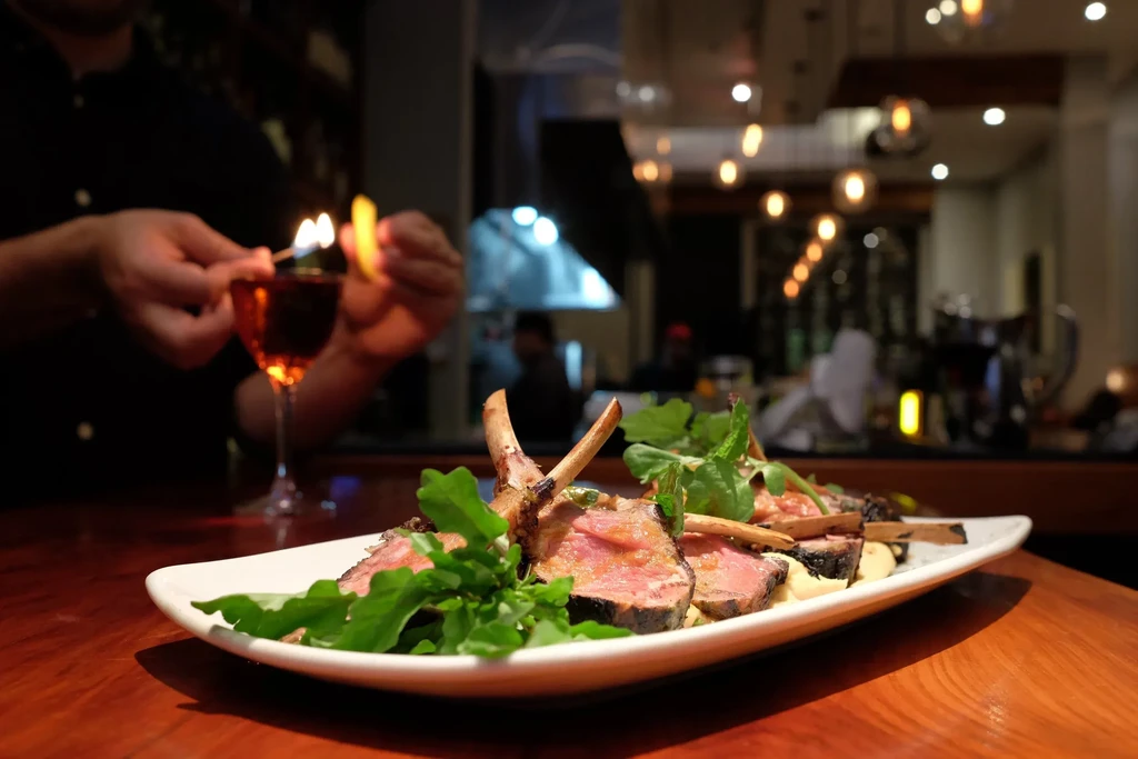 A bartender flaming an orange peel for a drink, with a plate of lamb chops.