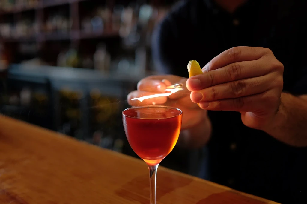 Bartender garnishes cocktail with burning citrus peel over glass on bar.