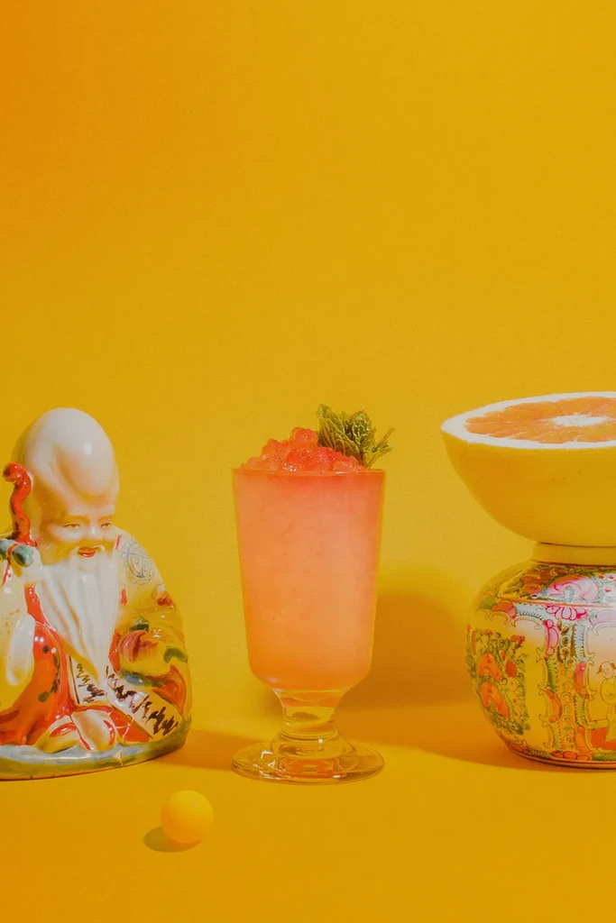 A yellow-orange cocktail, ceramic Buddha statue, and grapefruit on yellow background.
