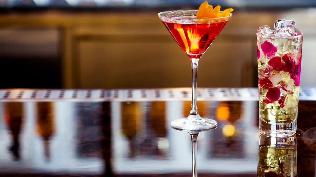 Two cocktails, one red and one with flower petals, on a bar.