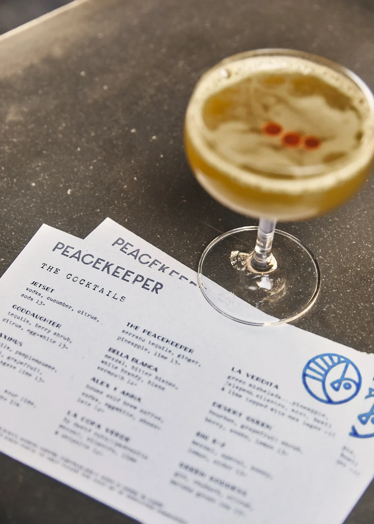 A cocktail glass and a "Peacekeeper" menu sit on a gray tabletop.
